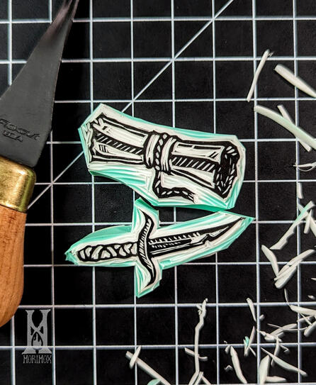 Blockprint stamp of a rolled up scroll with a sleeve and small rope securing it. Below it is a stamp of a small dagger with a wrapped handle. The stamps are resting on a black and white grid cutting mat with shavings of rubber scattered around them.
