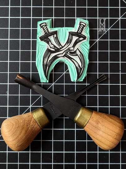 Blockprint stamp of two twin swords with the blades crossing each other. The flexcut gouges used to carve it are crossed in a similar manner below, mirroring the stamp.