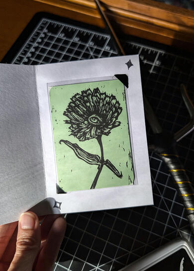 An Art Card Edition and Original (ACEO) trading card made with black ink on seafoam green paper. The print depicts a flower with an eye at the center replacing the disc florets, two small dewdrop tears on the petals, and leaves in a wistful pose.