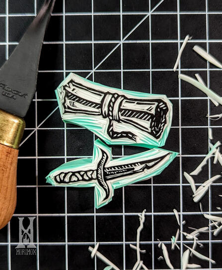 Blockprint stamp of a rolled up scroll with a sleeve and small rope securing it. Below it is a stamp of a small dagger with a wrapped handle. The stamps are resting on a black and white grid cutting mat with shavings of rubber scattered around them.