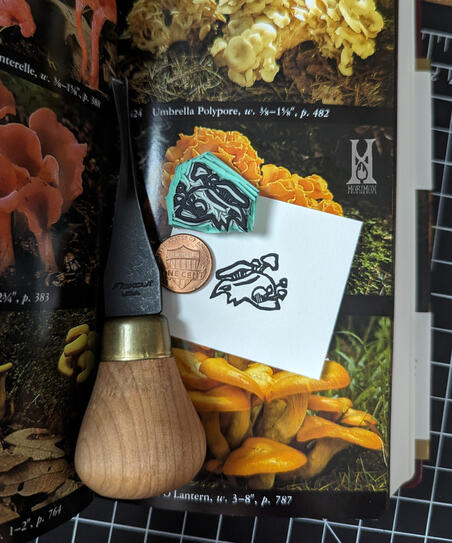 Linocut stamp and print of an animal skull with several mushrooms growing out of it. A penny is accompanying them for scale, showing that the stamp is just slightly larger than the coin. The stamp and print are resting on an open mycology book.