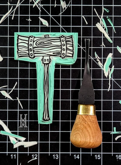 A blockprint of a mallet posed on a black and white grid cutting mat along with a flexcut gouge and shavings of linoleum framing it.