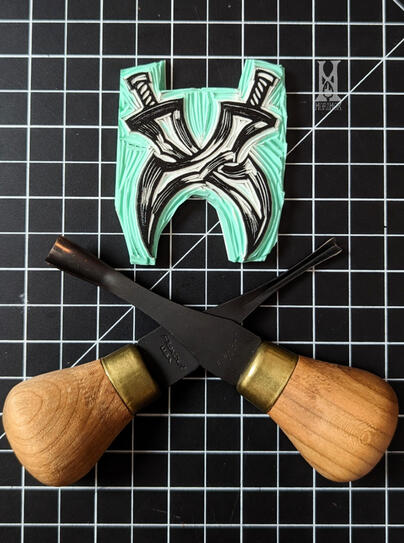 Blockprint stamp of two twin swords with the blades crossing each other. The flexcut gouges used to carve it are crossed in a similar manner below, mirroring the stamp.