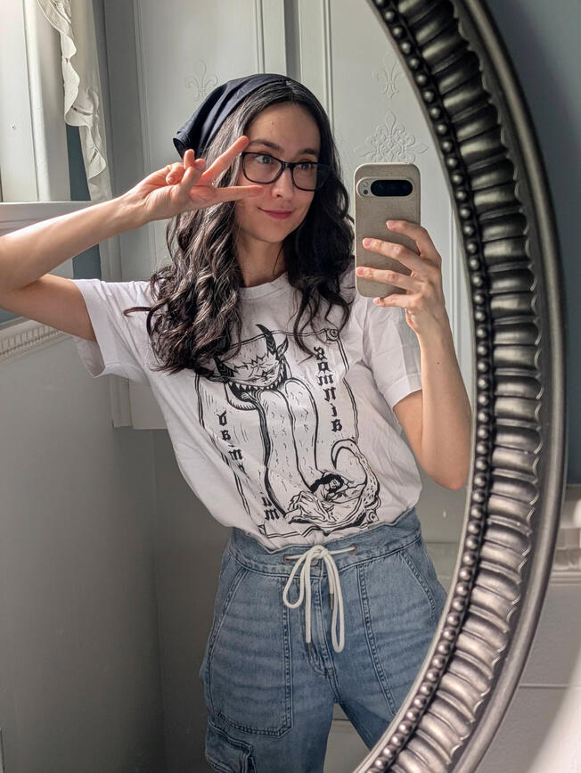 The artist Morimox A woman with below shoulder length brown curly hair poses in front of a mirror taking a selfie and giving a peace sign.