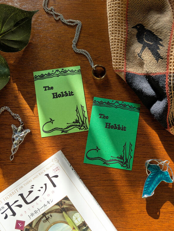 The Hobbit Mini Blockprint Two small blockprints of the a cover of The Hobbit books in light green and dark green variants. On the cover is a mountainside with a sun and moon at the top and a dragon on the bottom. The prints are decoratively surrounded by Lord of the Rings jewelry,